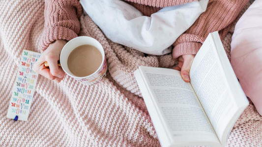 Person holding a mug and an open book on a pink blanket